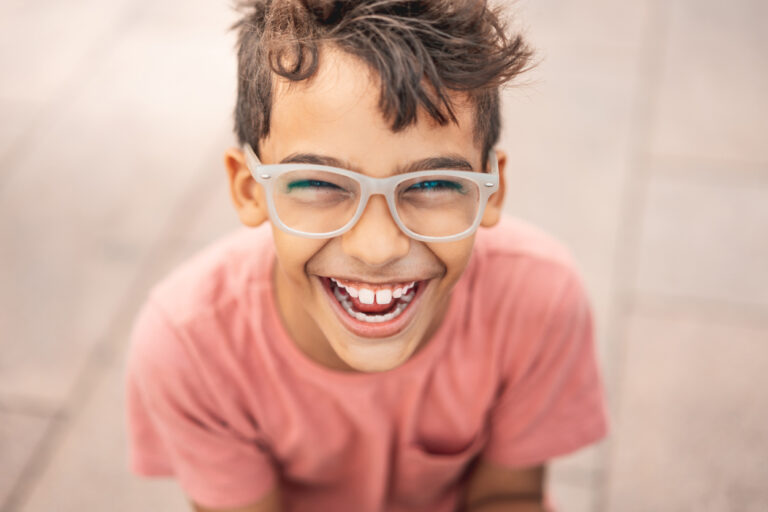 Young boy wearing salmon colored t-shirt and gray glasses smiling