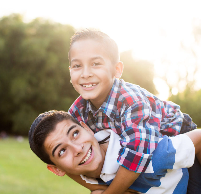 A latin teen boy carrying his younger brother on the back and smiling in a horizontal waist up shot outdoors.