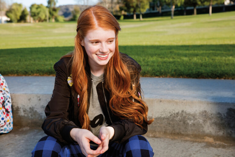 Ginger teen girl sitting on concrete at park smiling