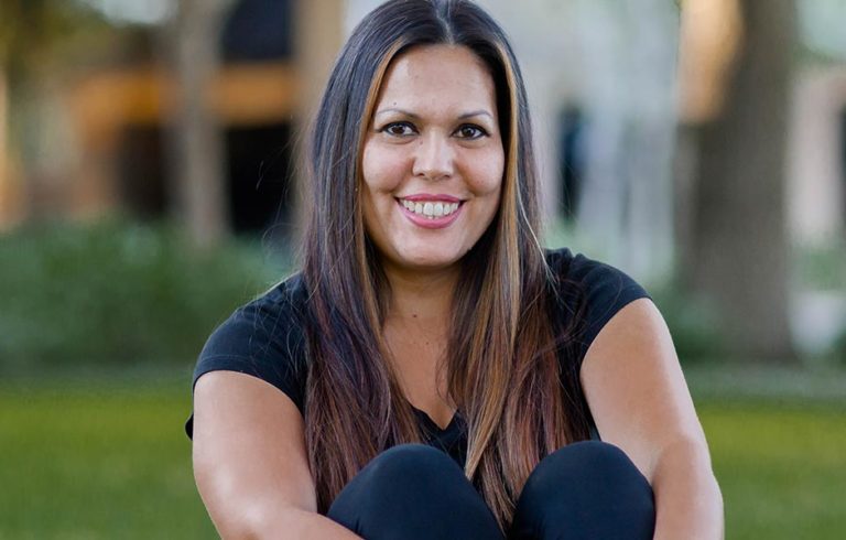 Latina woman wearing black t-shirt and pants sitting in grass with knees up smiling