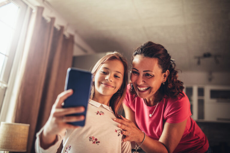Older woman wearing pink shirt standing with young girl wearing white top and holding smartphone in living room